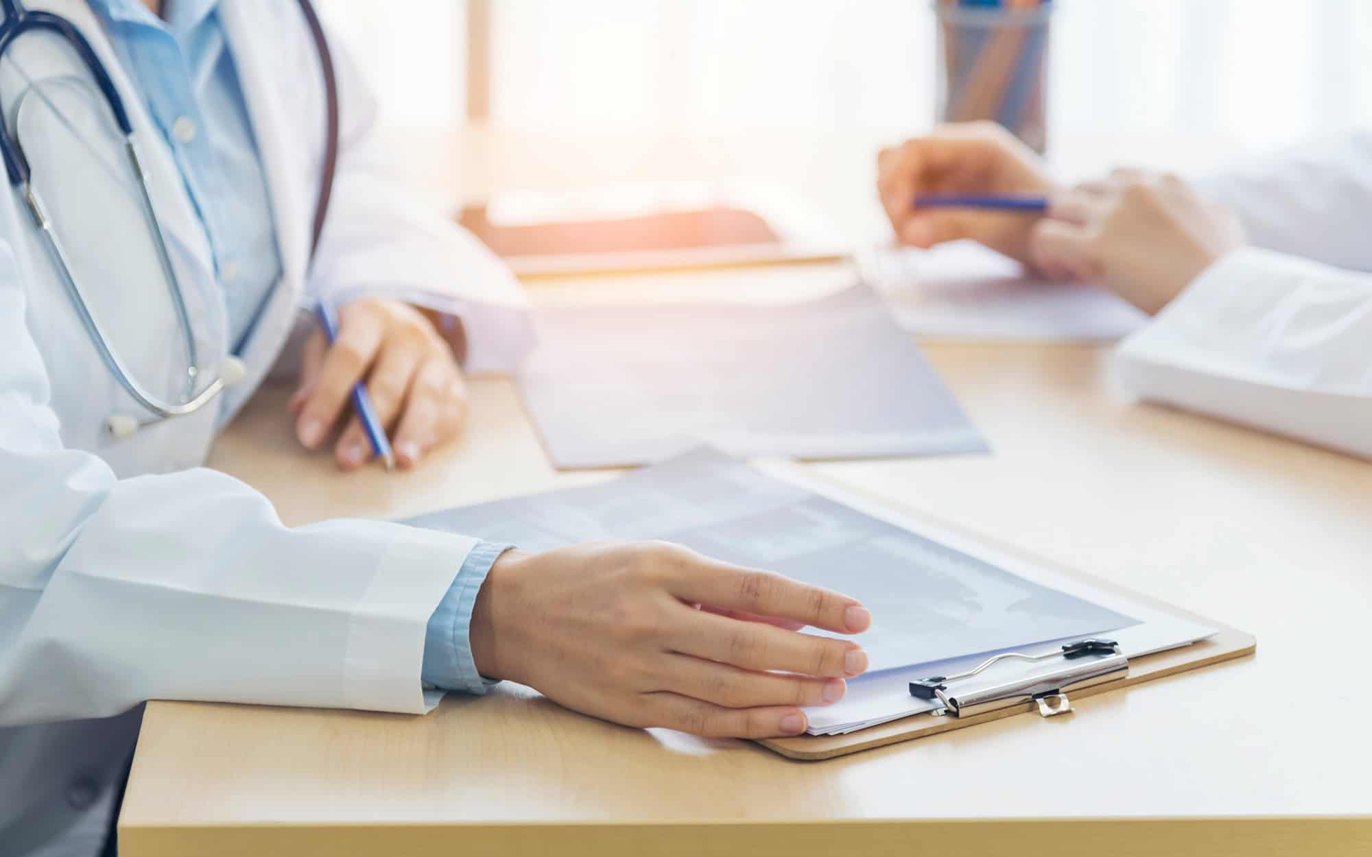Side View Of A Doctor's Hand Laying Next To Clipboard In Meeting